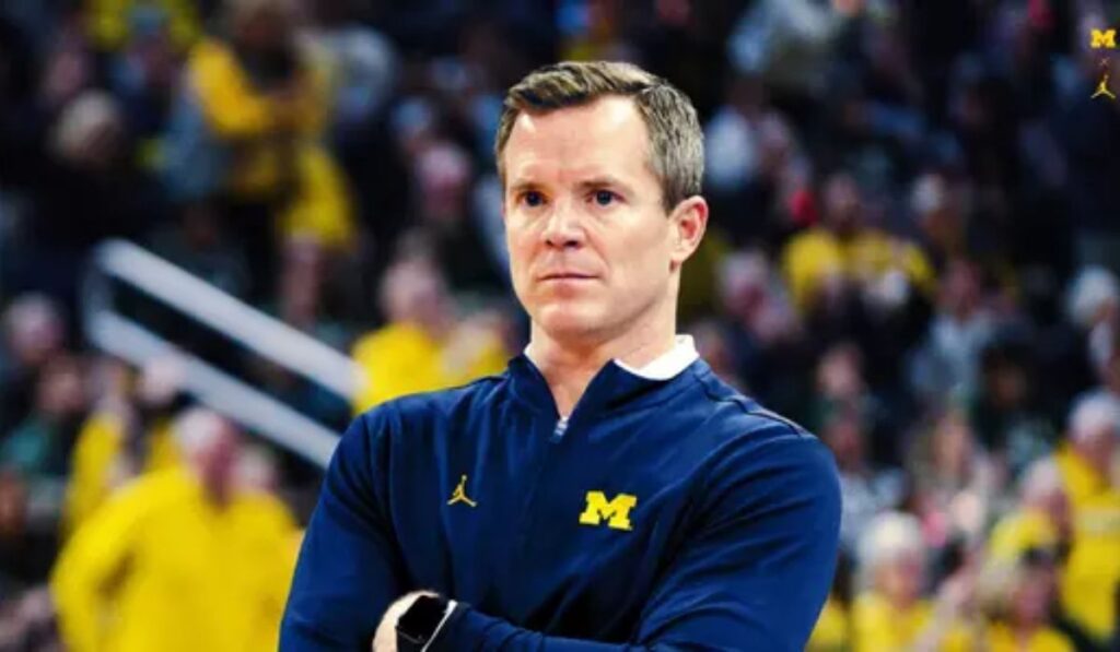 Image of Michigan coach Dusty May during a post-game interview, sharing insights about the team's victory, with players in the background celebrating, a scoreboard reflecting the win, and reporters capturing the moment.