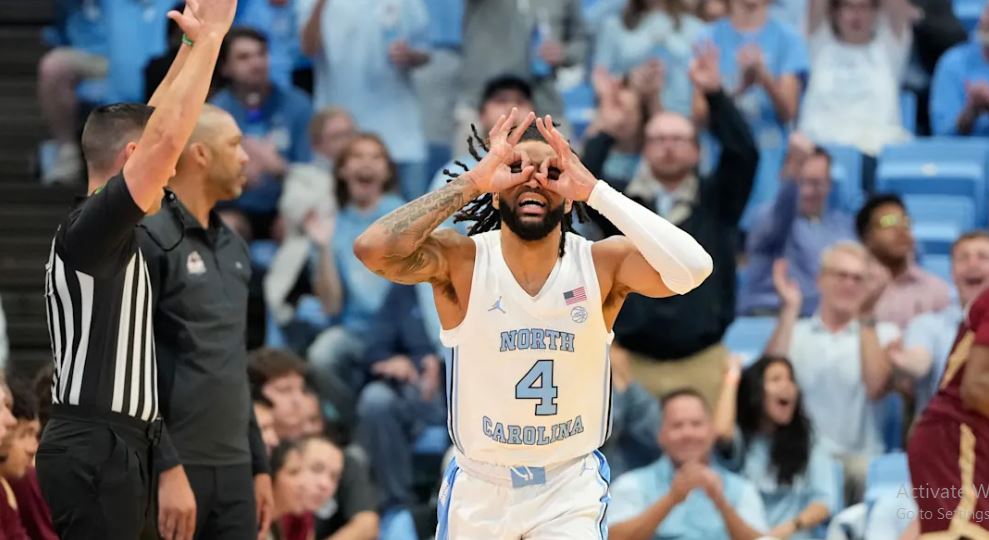 Action shot of the North Carolina Tar Heels basketball team celebrating a close victory over Elon, with players embracing on the court, fans in the stands erupting with excitement, and the scoreboard indicating a thrilling finish.