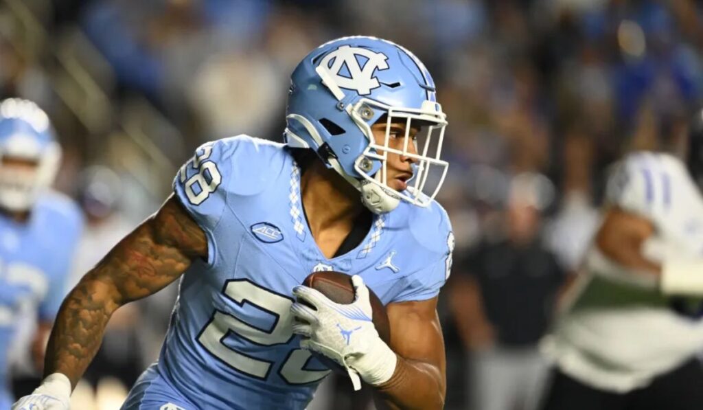 Omarion Hampton, UNC running back, celebrates on the field after achieving 3,000 career rushing yards, surrounded by teammates and fans cheering in the background.