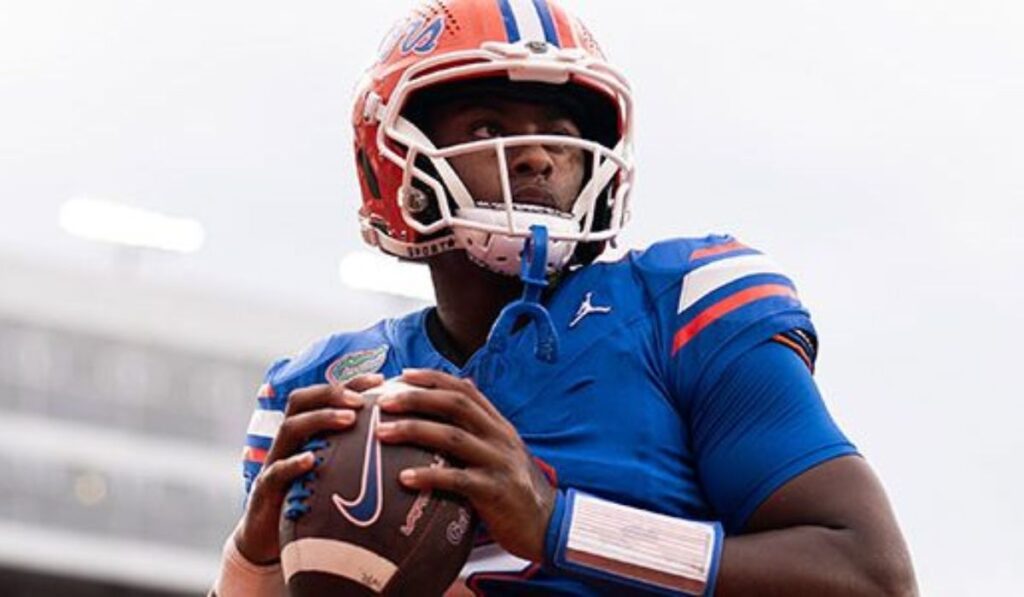 Image of DJ Lagway, Florida Gators quarterback, smiling during a press conference, with a backdrop featuring the Gators logo and sports reporters in attendance, reflecting the positive update on his recent hamstring injury.